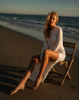 The Shea Long Sleeve T-Shirt Dress in White — seated on the shoreline showing the drape and high slit